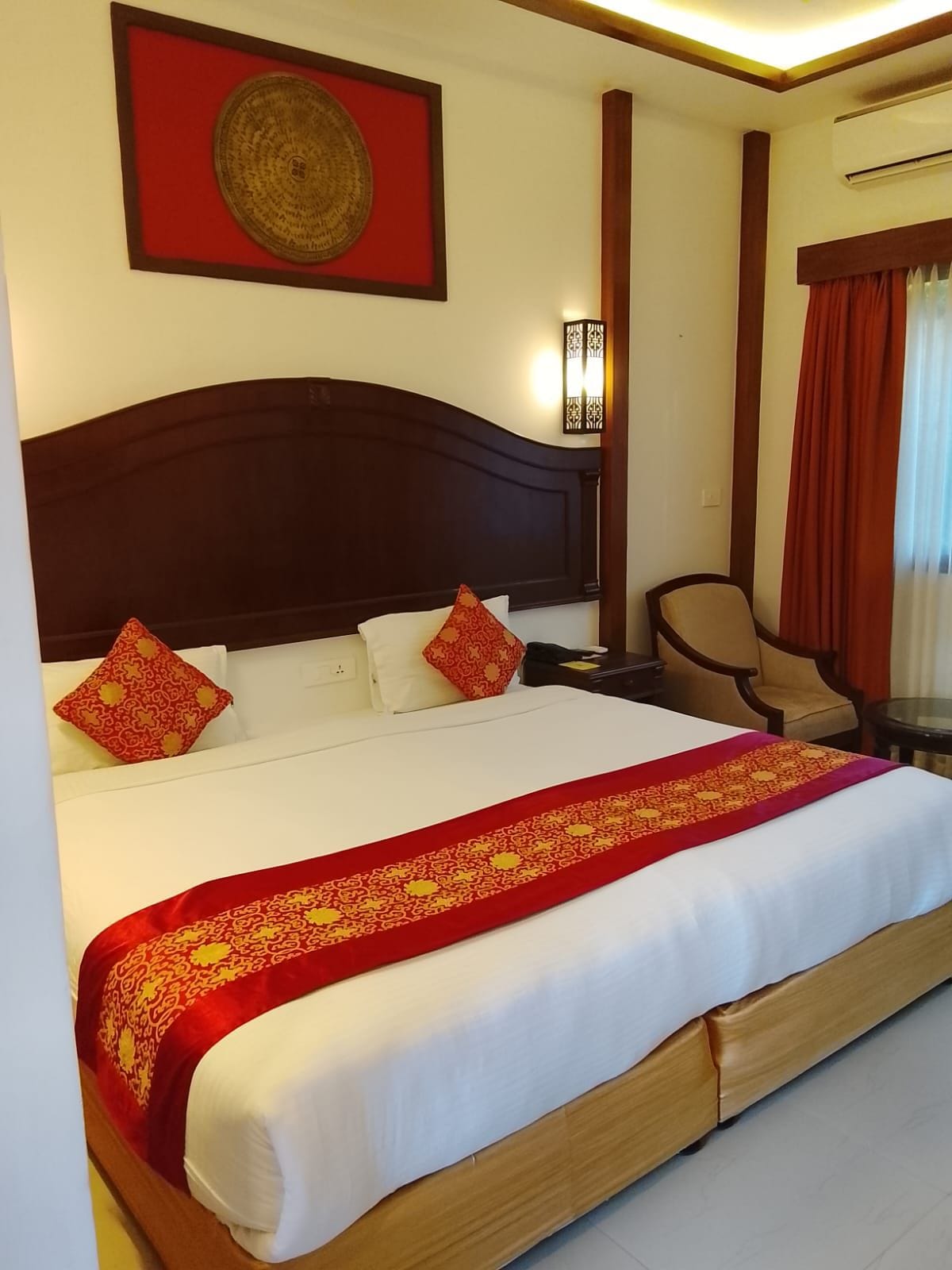 A hotel room with a large bed, a headboard, a nightstand, a chair, a table, and a wall-mounted lamp.