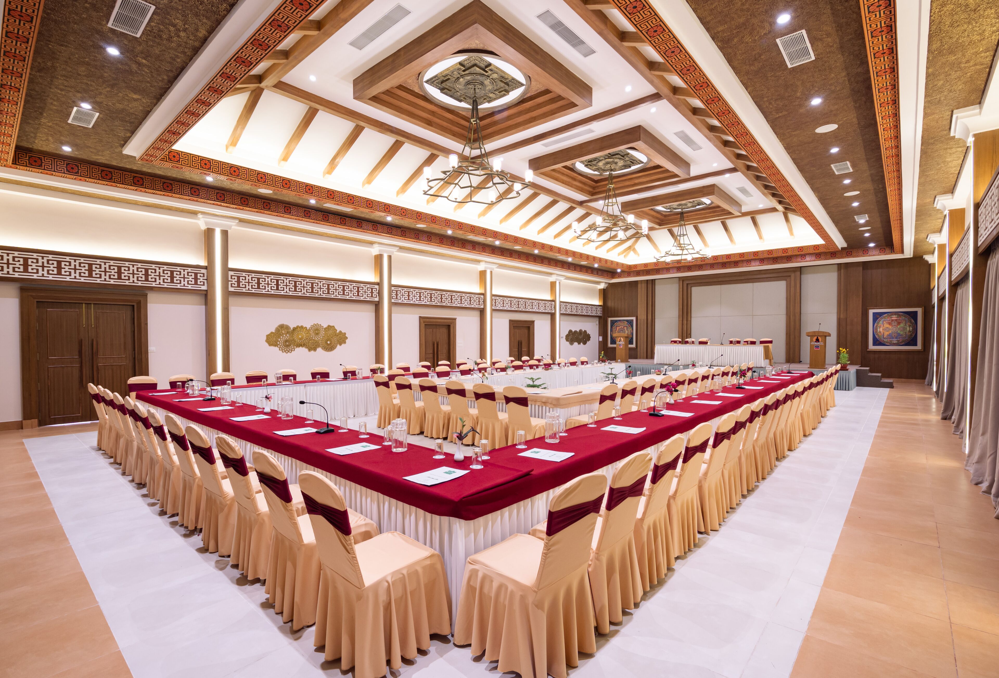 Un amplio salón de banquetes con una larga mesa dispuesta para un evento formal, que cuenta con un techo alto con intrincados trabajos de madera y una lámpara moderna.