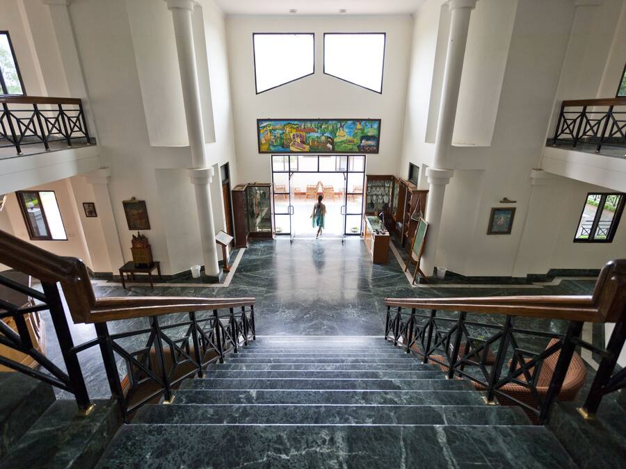 Una gran escalera con escalones de mármol y barandillas de madera conduce a un amplio vestíbulo con un gran ventanal y un mural colorido.