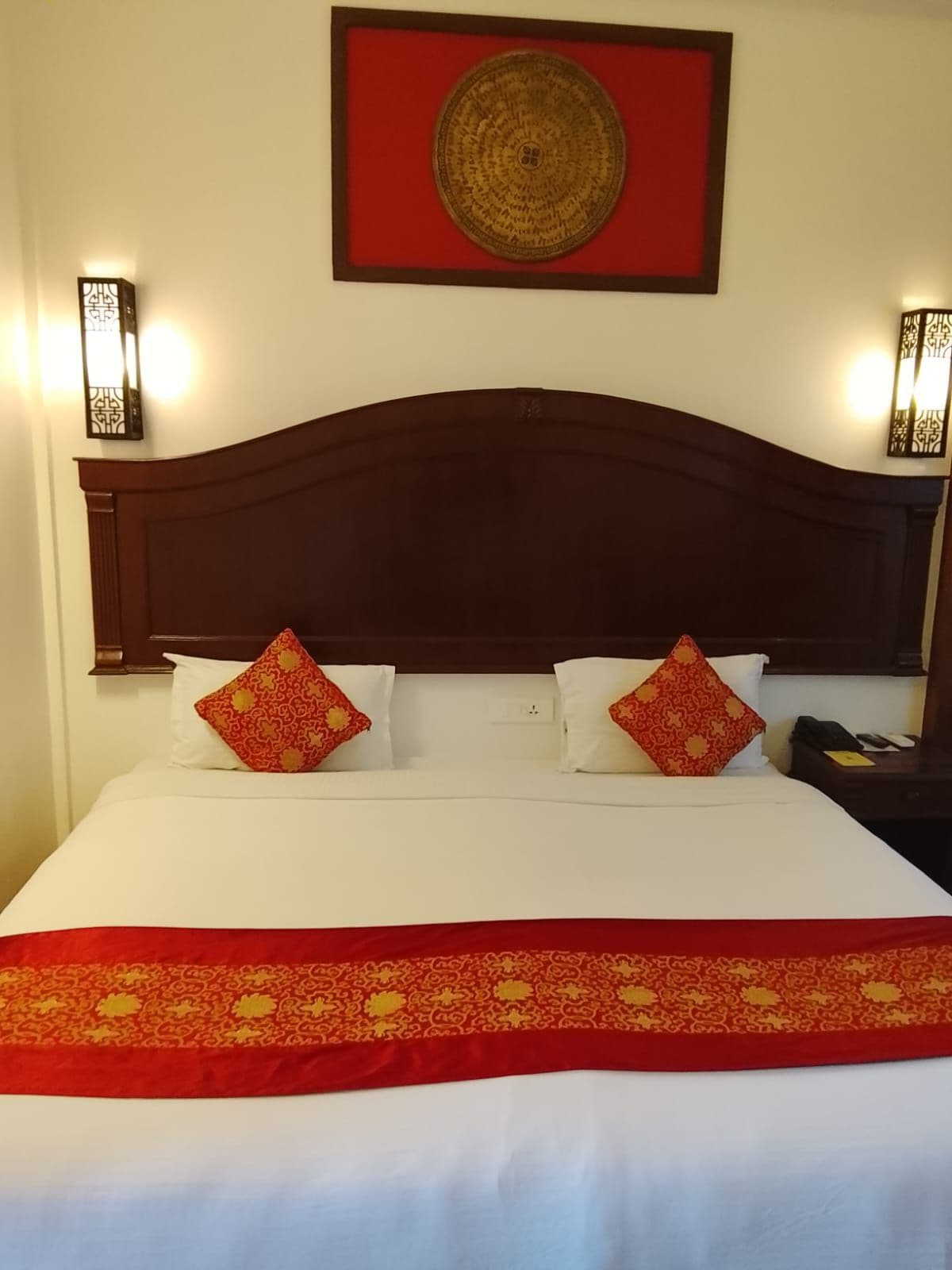 A hotel room with a large bed, two bedside lamps, a decorative wall hanging, and a red and gold bedspread.