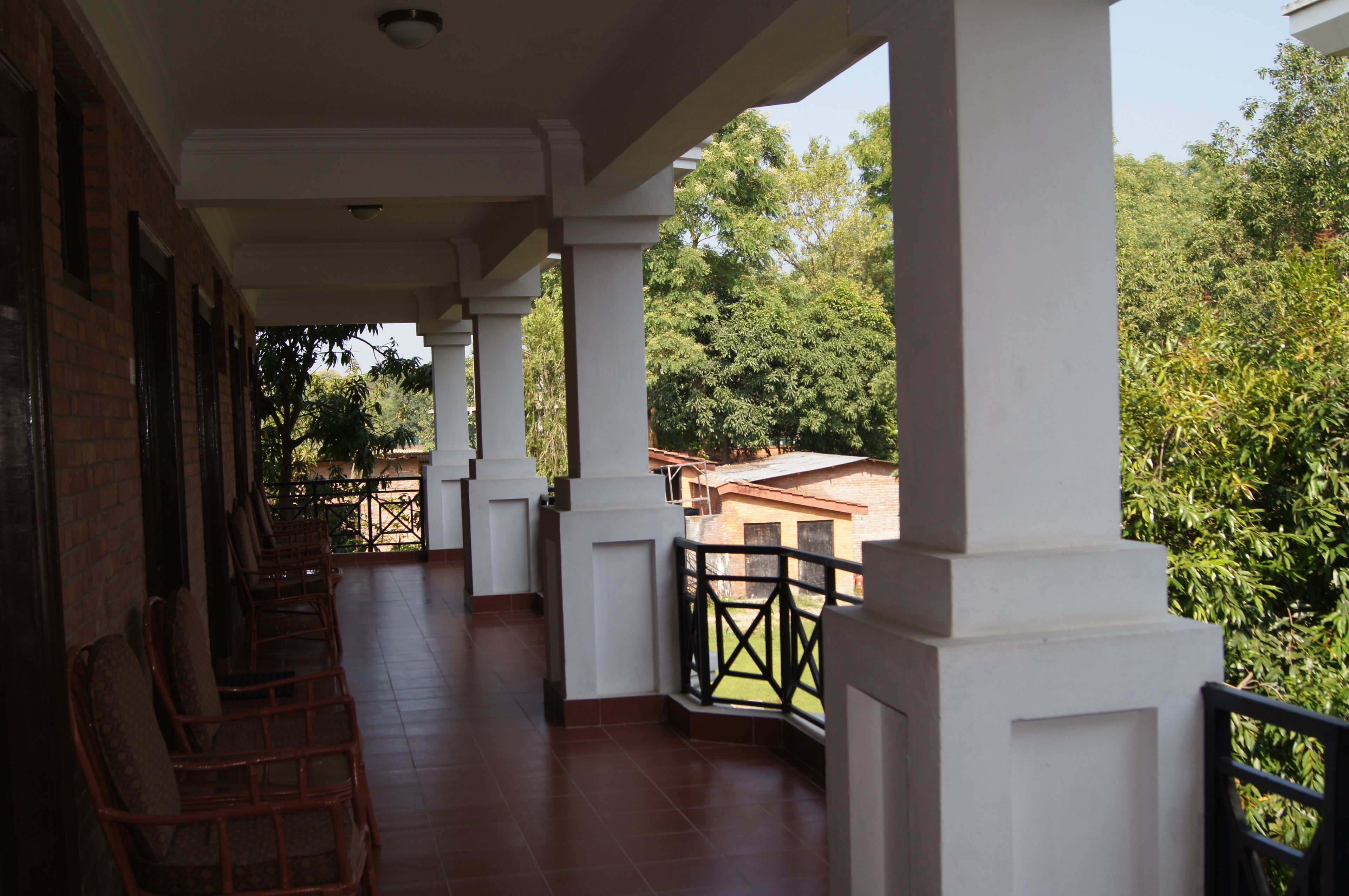 Un espacio al aire libre cubierto con columnas blancas, piso de baldosas y barandilla con vistas a un jardín.