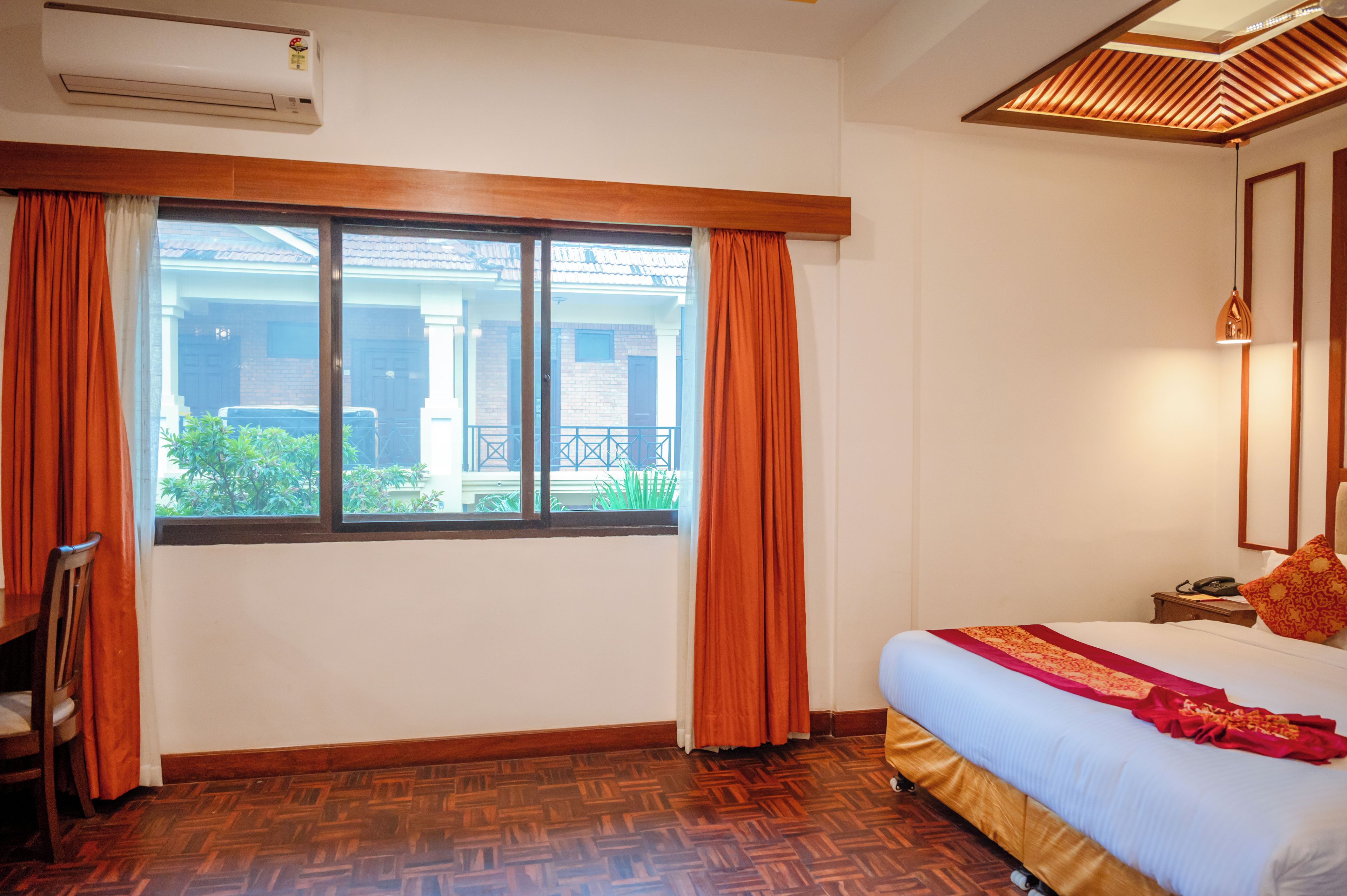 A hotel room with a large window, wooden flooring, a bed with a red and white bedspread, and a nightstand with a phone.