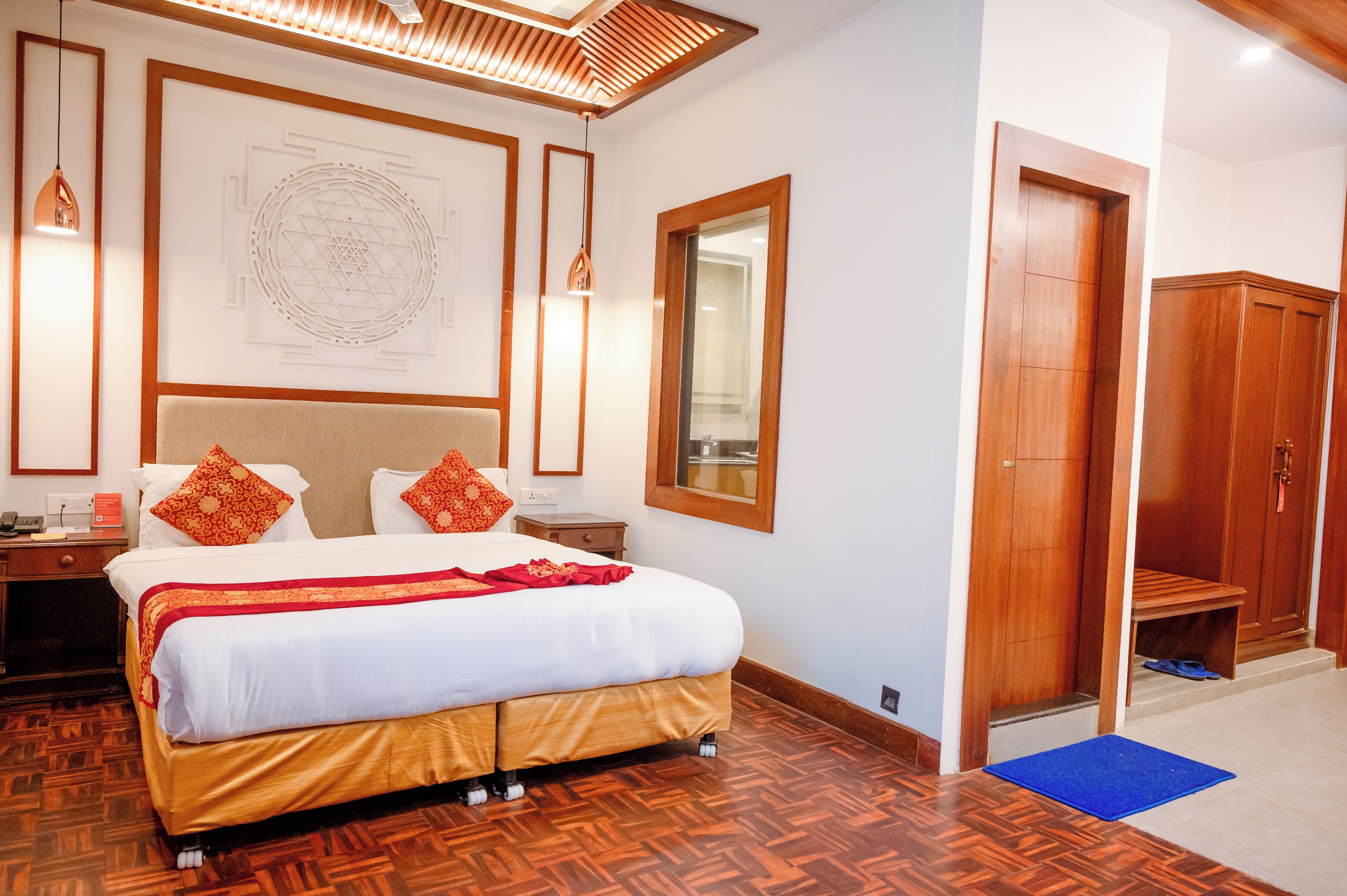 Habitación de hotel con una cama grande, suelo de madera, techo con una lámpara geométrica y un armario de madera, además de un felpudo azul en la entrada.