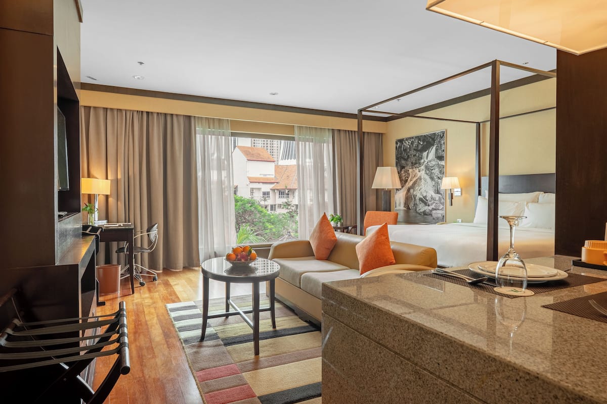 A hotel room with a bed, a desk, a sofa, a dining area, and a view of buildings outside.