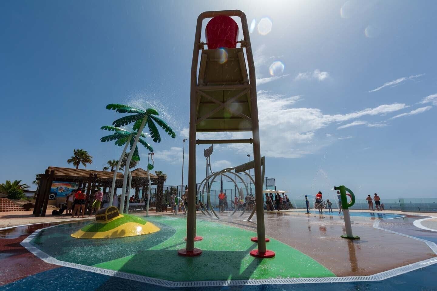 A water play area with a slide and splash pad, surrounded by palm trees and a beach view.
