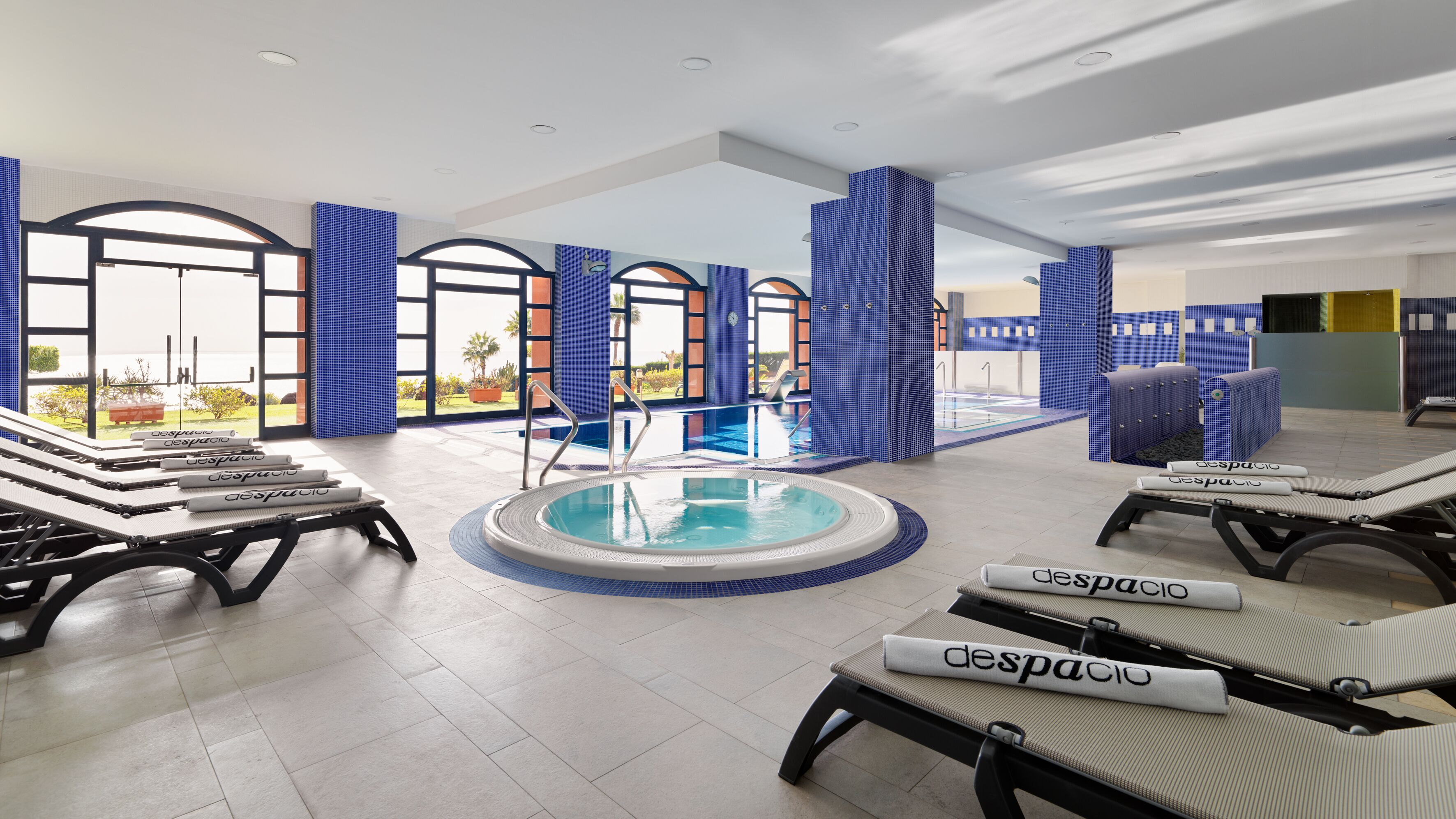Un espace piscine intérieur moderne, doté de chaises longues, d’un jacuzzi et de grandes fenêtres offrant une vue sur l’extérieur.