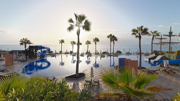 A pool area with lounge chairs and palm trees, surrounded by a wooden deck and a water slide. - H10 Tindaya (Pajara)