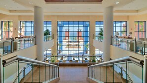 A spacious atrium with a central open area, glass railings, and a view of an outdoor plaza with palm trees and a red building. - H10 Tindaya (Pajara)