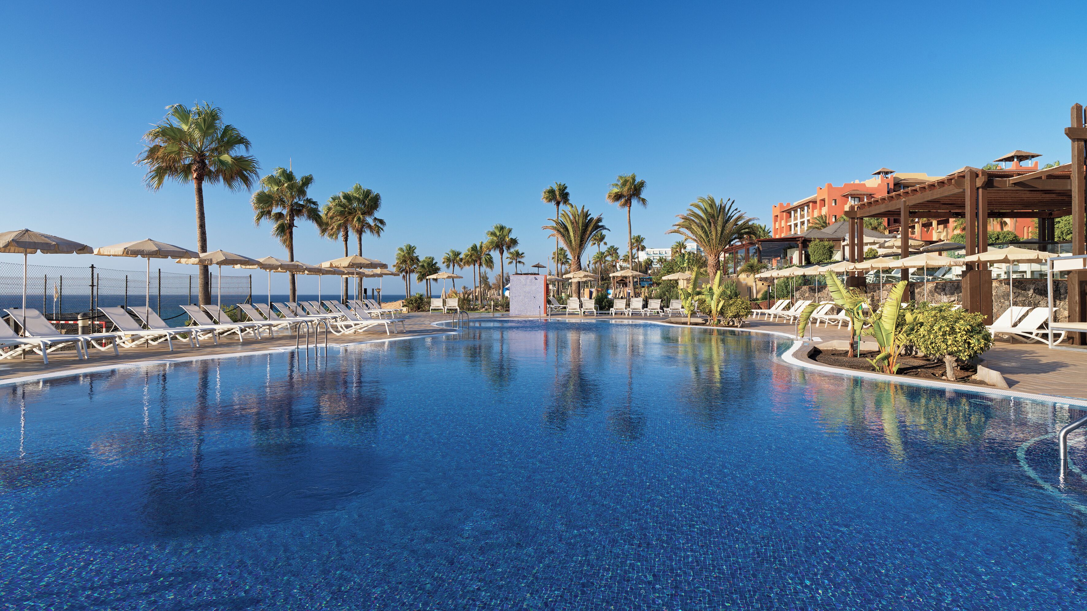 Ein Pool mit Liegestühlen und Sonnenschirmen, umgeben von Palmen und einem Gebäude mit roter Fassade.