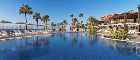 Ein Pool mit Liegestühlen und Sonnenschirmen, umgeben von Palmen und einem Gebäude mit roter Fassade.