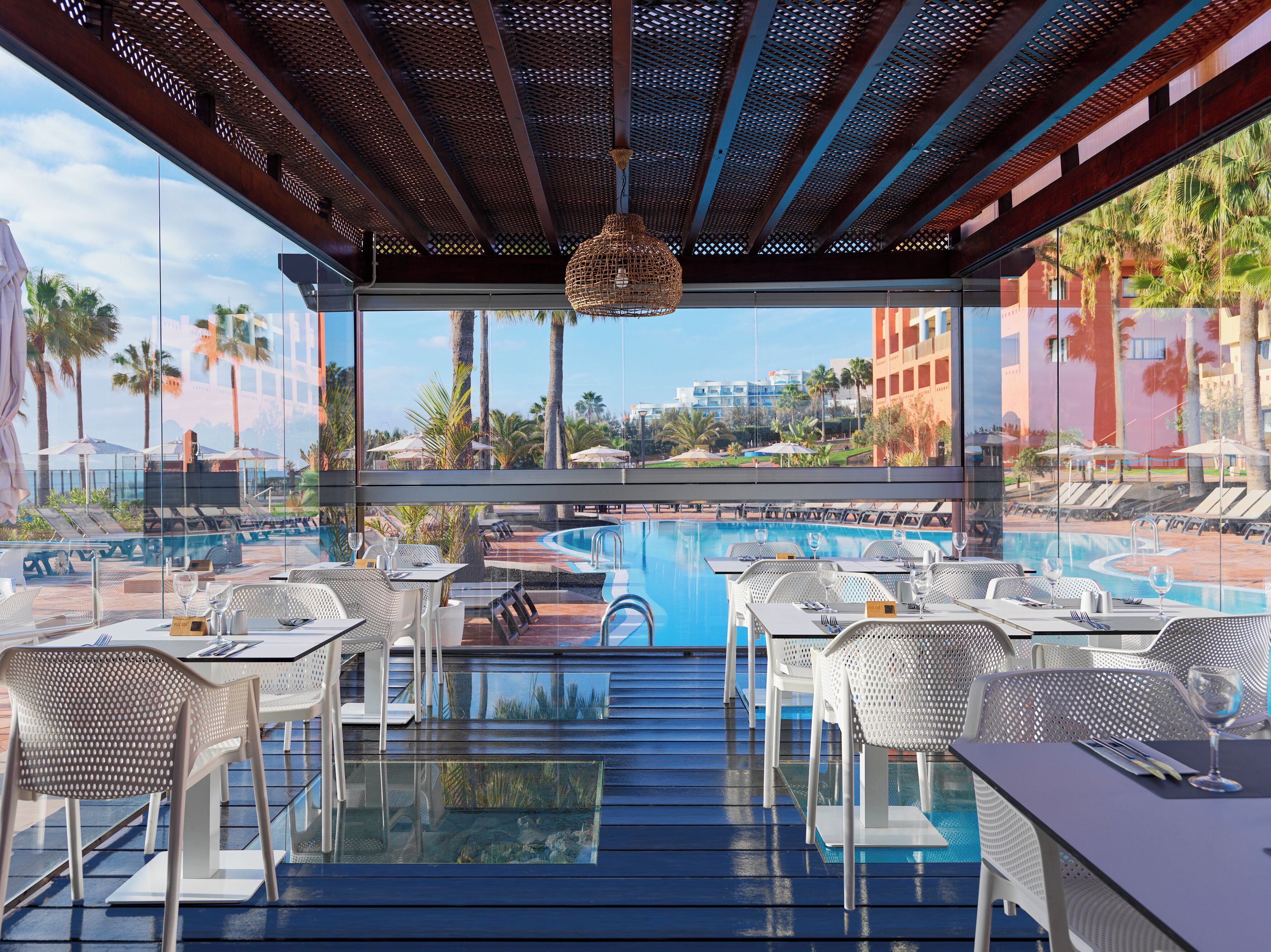 Ein Poolrestaurant mit Außenbereich, Blick auf einen Swimmingpool und Palmen.