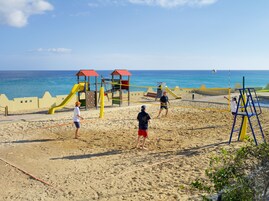 A beach volleyball court with a net, sand, and a playground with slides in the background.