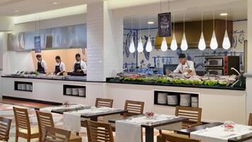 Un restaurante de mariscos con una barra que exhibe pescado fresco, una zona de cocina con cocineros y mesas preparadas para comer.