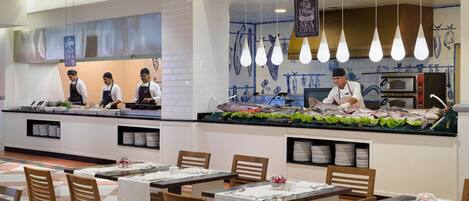 A seafood restaurant with a counter displaying fresh fish, a kitchen area with chefs, and tables set for dining.