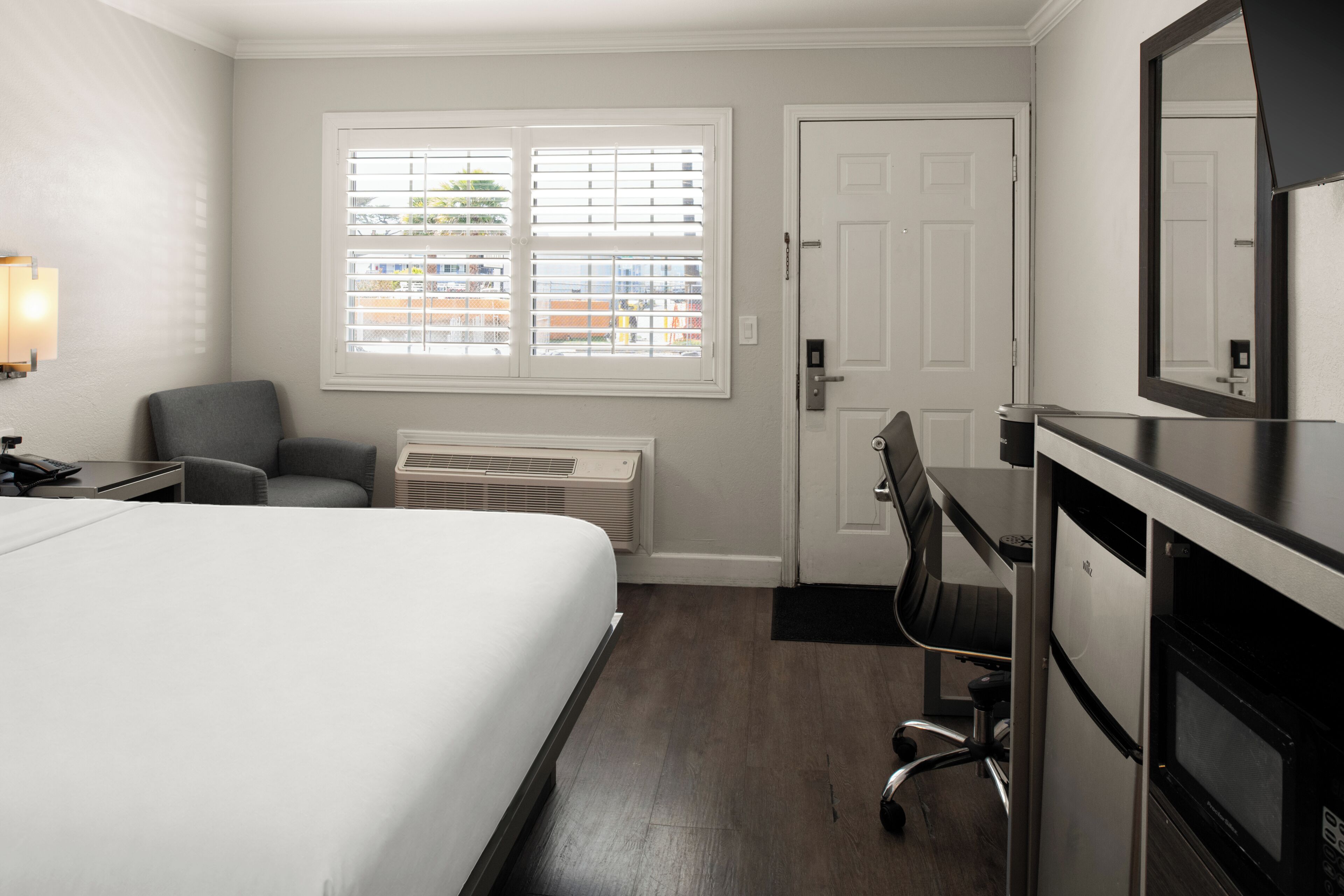 A hotel room with a bed, a desk, a chair, a radiator, a door, and a window with blinds.