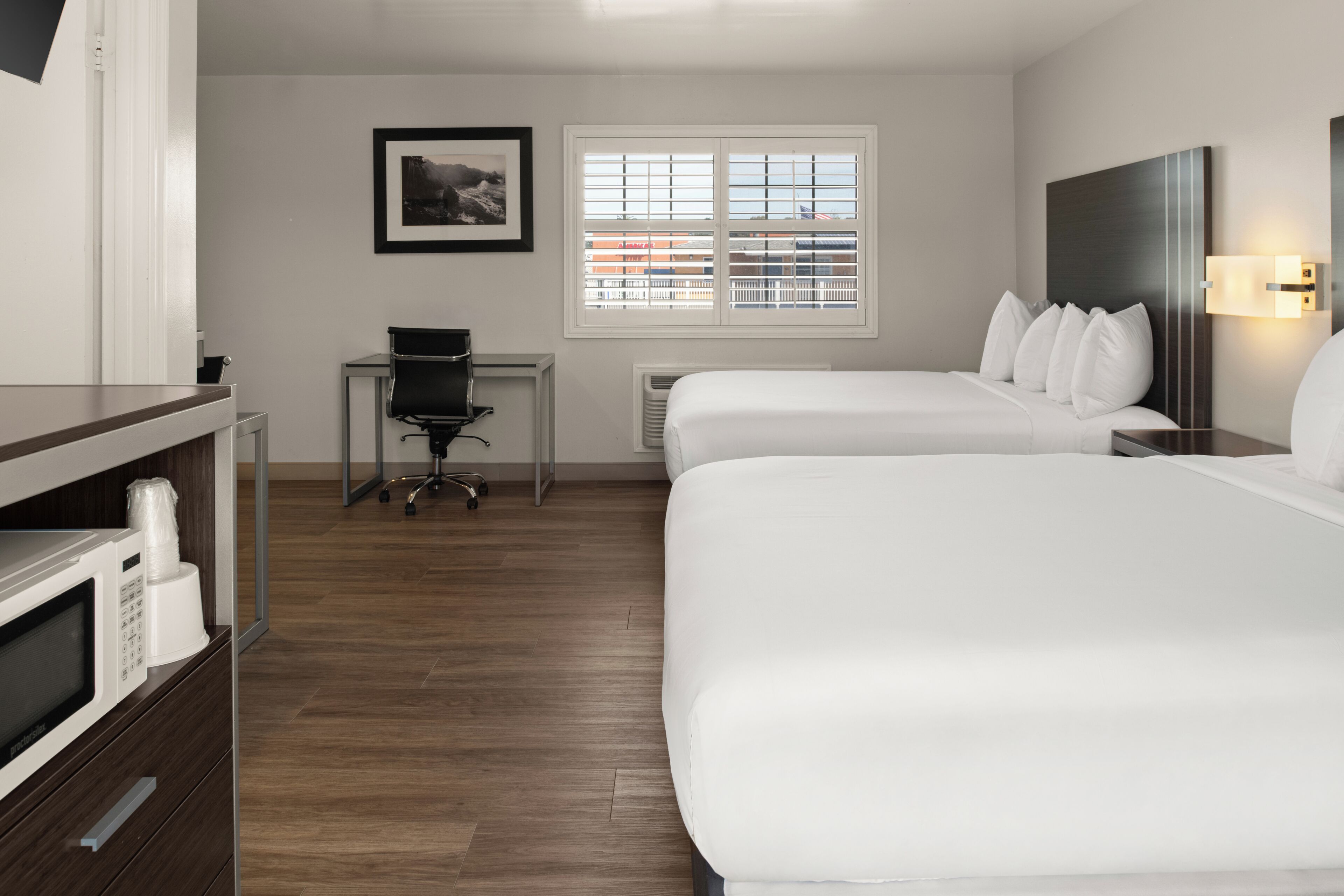 A hotel room with two beds, a desk, a chair, a microwave, and a framed picture on the wall.