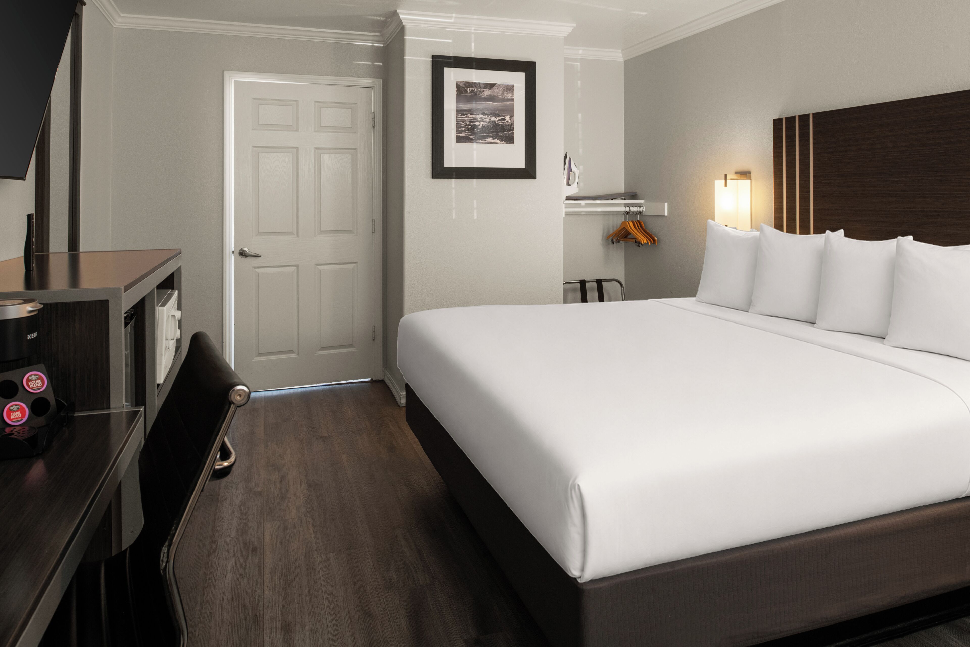 Habitación de hotel con cama, escritorio, silla, puerta, cuadro en la pared y perchero con ganchos.