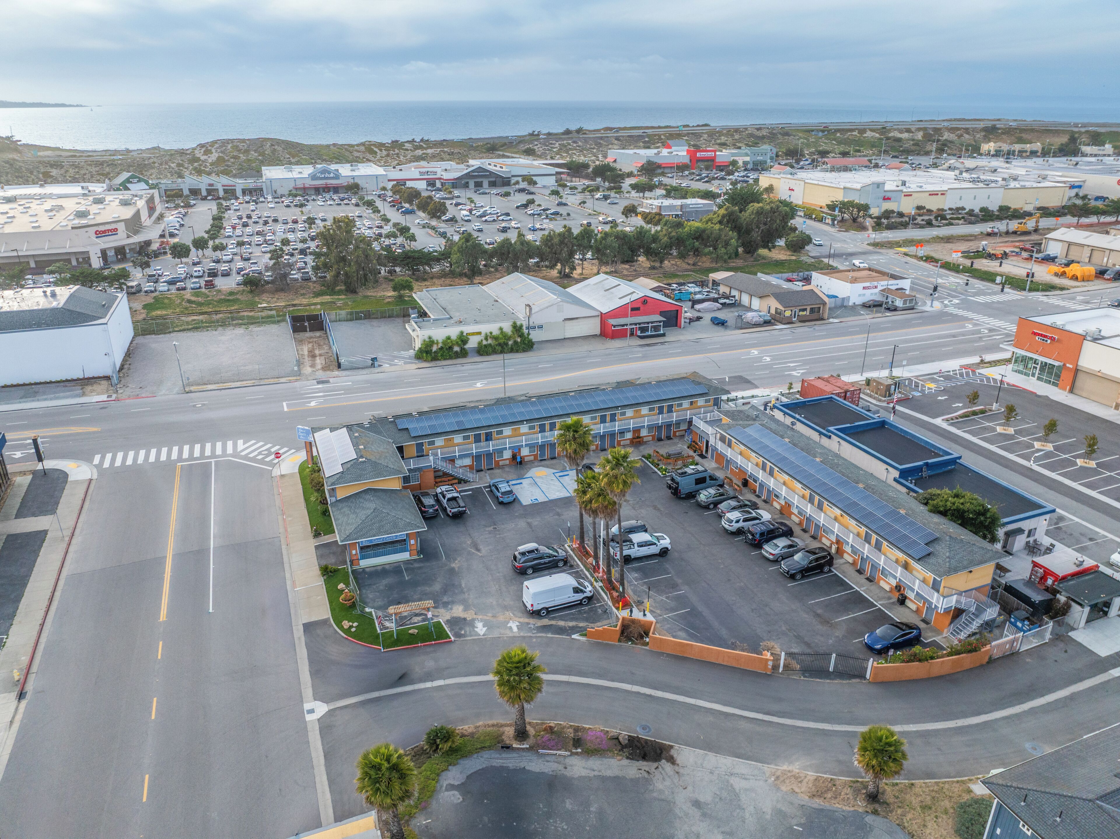 An aerial view of a commercial area with a parking lot, a building with solar panels, and a coastal backdrop.