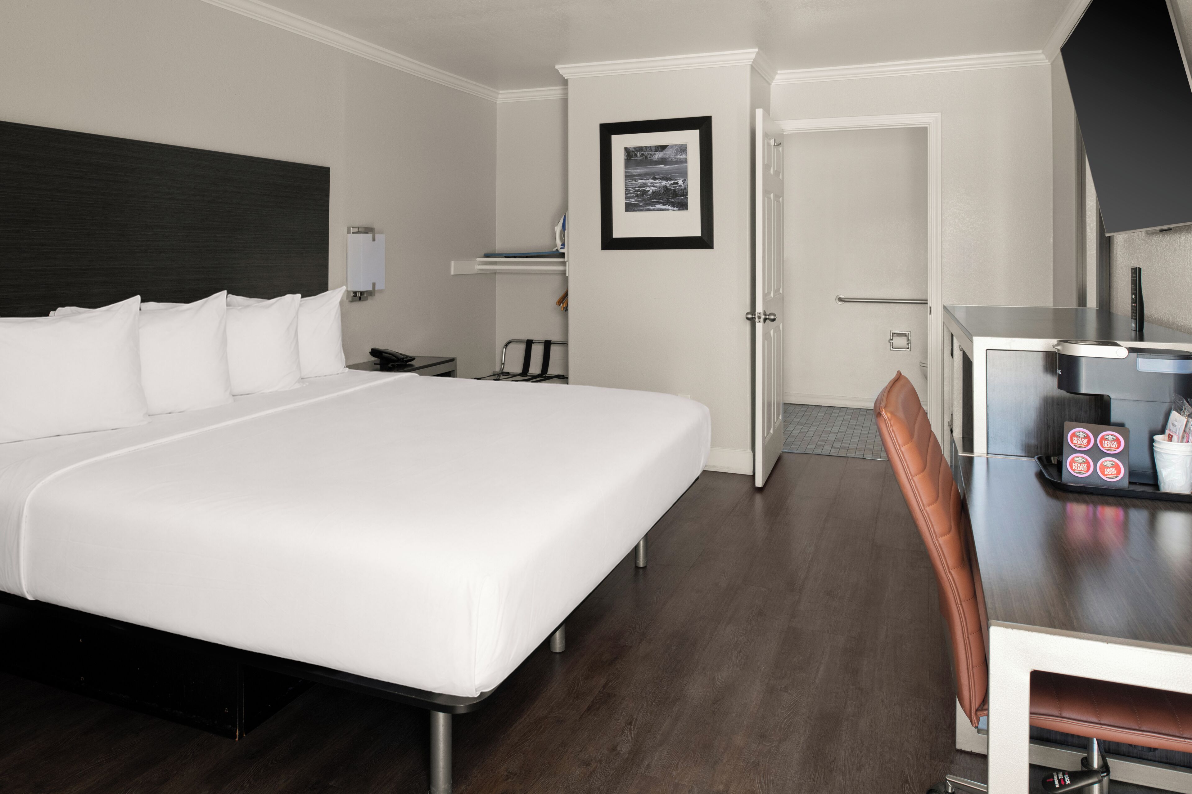 Habitación de hotel con una cama grande, un escritorio con silla, una mesita con cafetera y un cuadro en la pared.