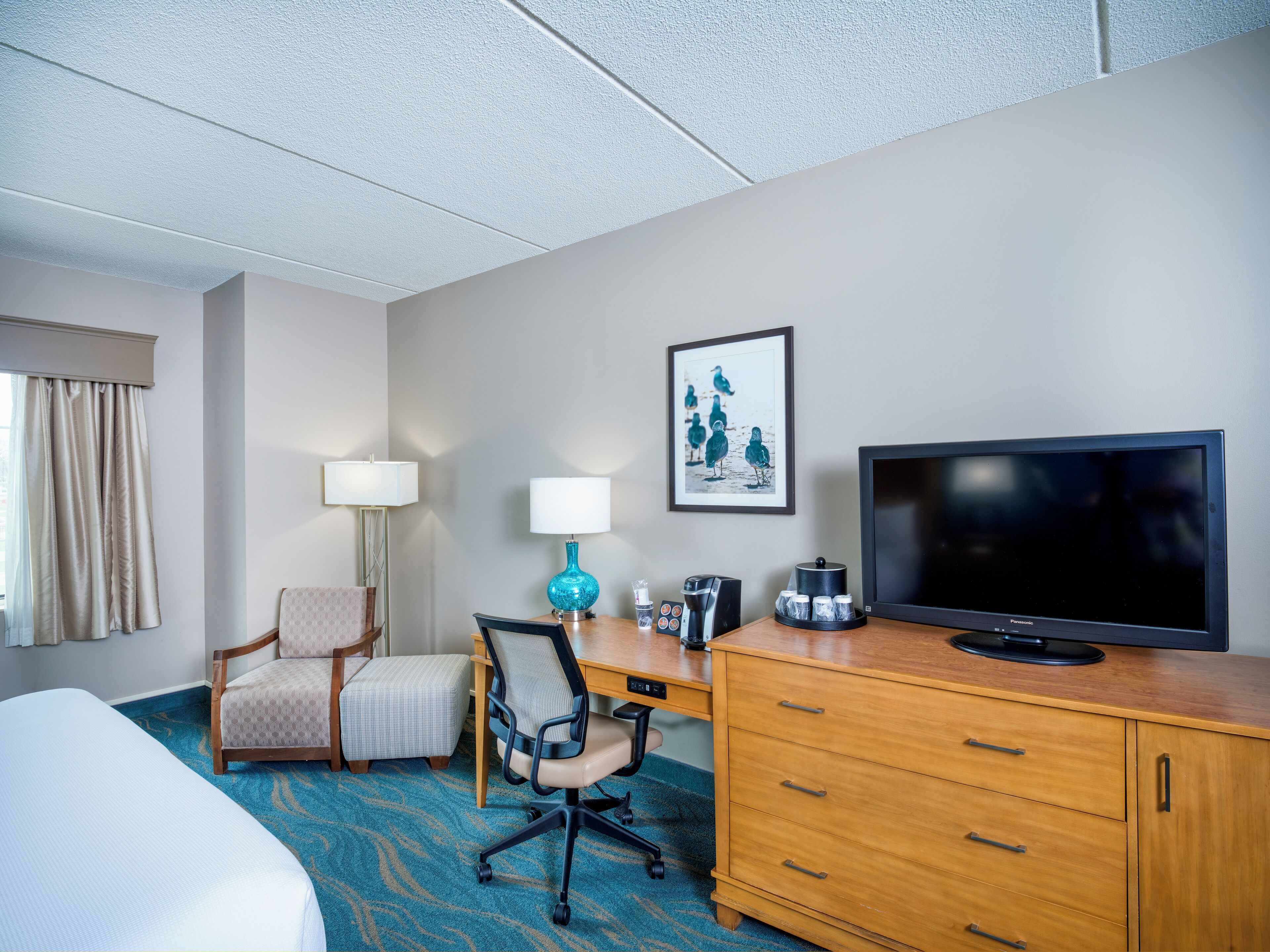 A hotel room with a bed, a desk with a chair, a television, a lamp, and a framed picture on the wall.