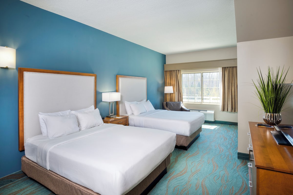 A hotel room with two beds, a desk, a chair, a lamp, a potted plant, and a window with curtains.