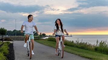 Two people riding bicycles on a paved path with a body of water and a cloudy sky in the background.