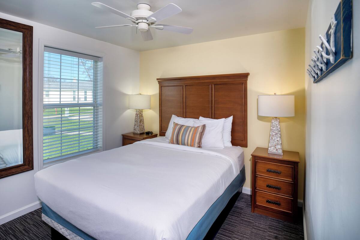 A hotel room with a large bed, wooden headboard, two bedside tables with lamps, a ceiling fan, and a window with blinds.