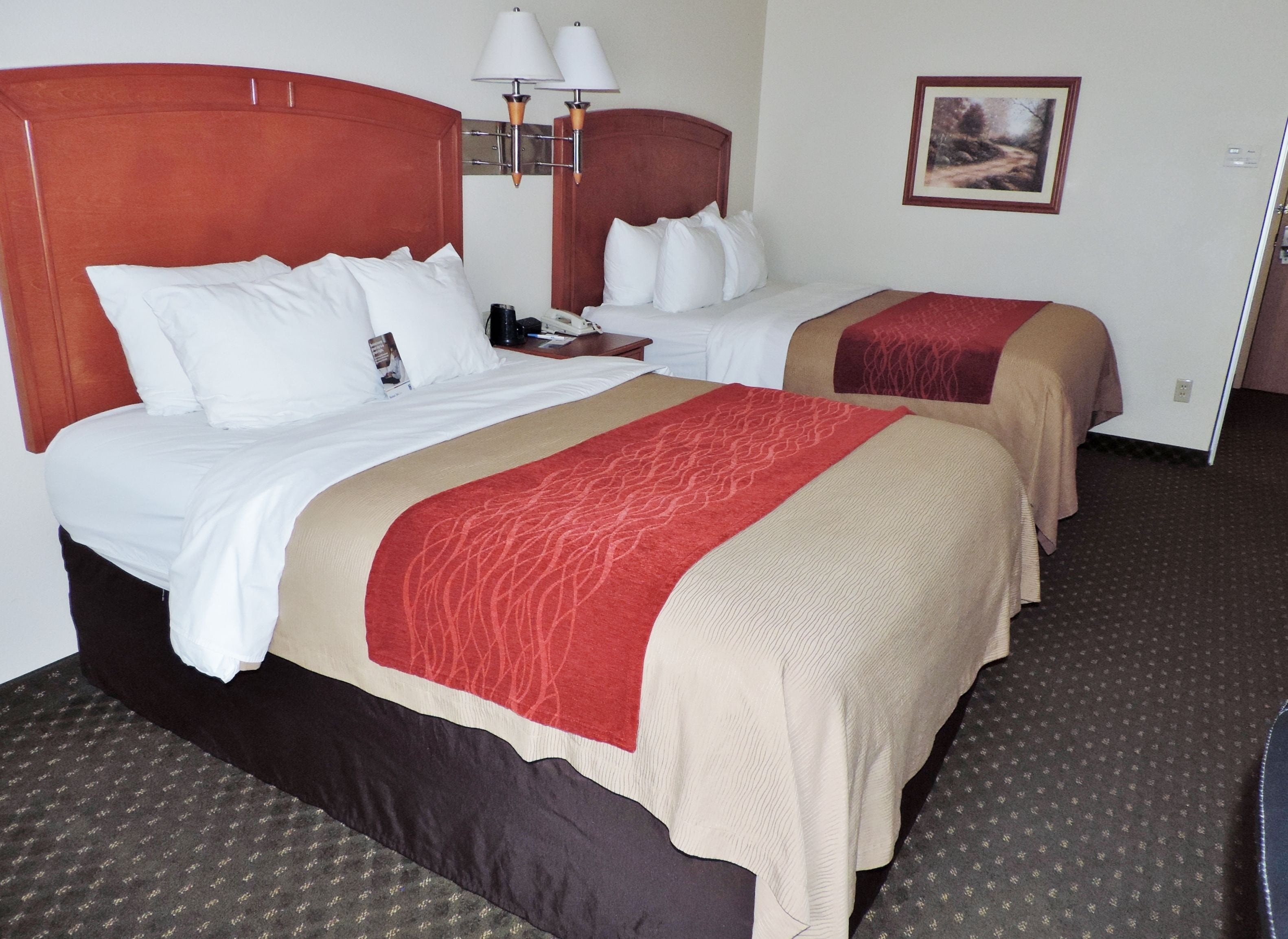 A hotel room with two beds, each with white bedding and red and beige throw blankets. There are two wall-mounted lamps above the beds and a framed picture on the wall.