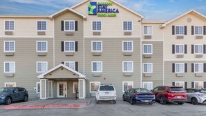 Un hotel de varios pisos con un letrero que dice "extended stay America select suites". Hay varios coches estacionados al frente, y el edificio tiene una fachada simétrica con ventanas y unidades de aire acondicionado uniformemente distribuidas.