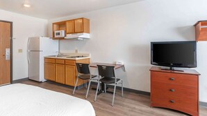 Habitación de hotel compacta con kitchenette, una cama, un escritorio con silla y un televisor sobre un aparador de madera.