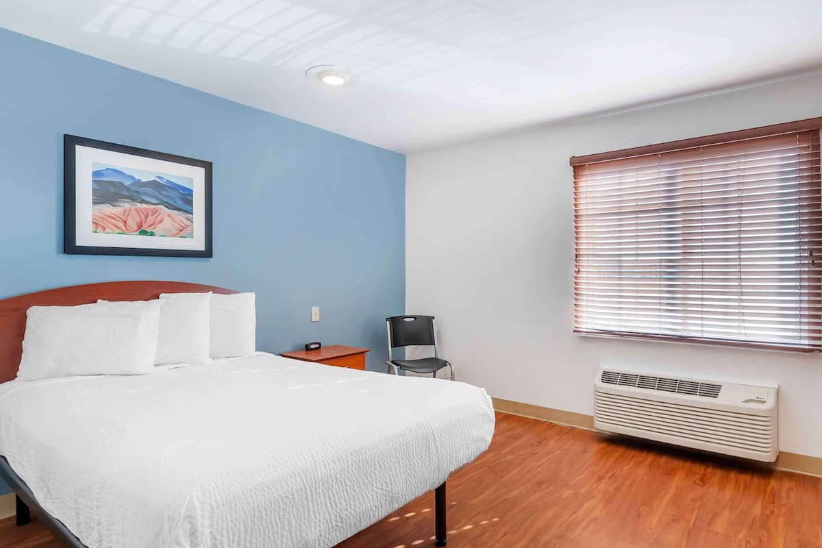 Um quarto com cama, uma cadeira, aquecedor, janela com persianas e um quadro pendurado na parede.