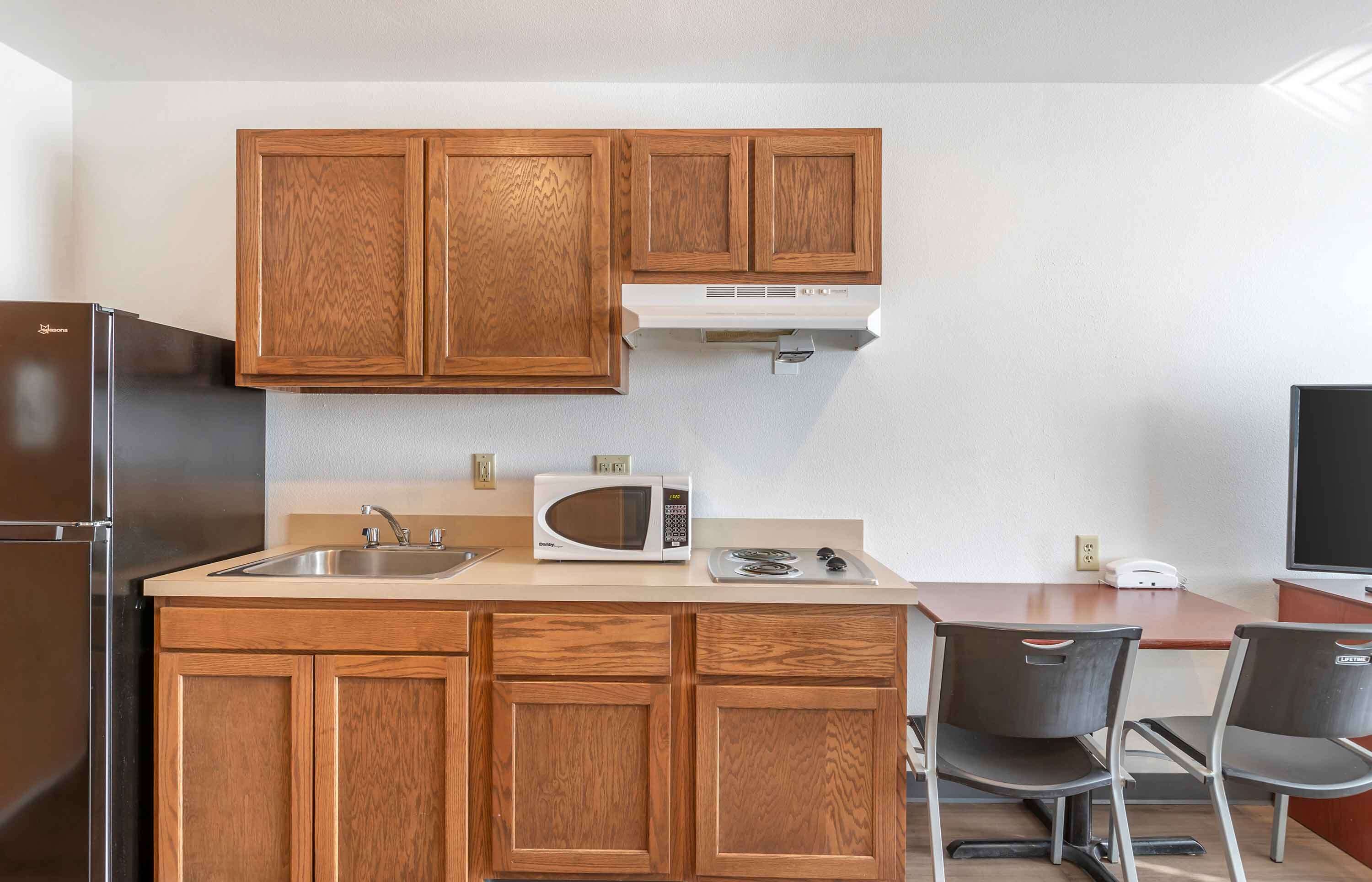 Una cocina compacta con gabinetes de madera, un refrigerador de acero inoxidable, un microondas y un pequeño comedor con dos sillas.