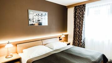 Habitación de hotel con cama, lámparas de noche, mesita de noche, un cuadro colgado en la pared y una ventana con cortinas estampadas.