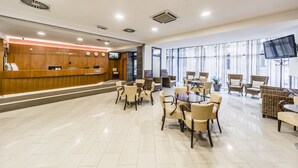 A hotel reception area with a wooden reception desk, a seating area with wicker furniture, and large windows providing natural light.