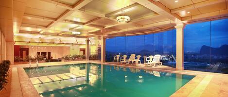 Una piscina interior con agua azul cristalina, rodeada de sillones de descanso y un amplio ventanal con vistas a la ciudad al atardecer.