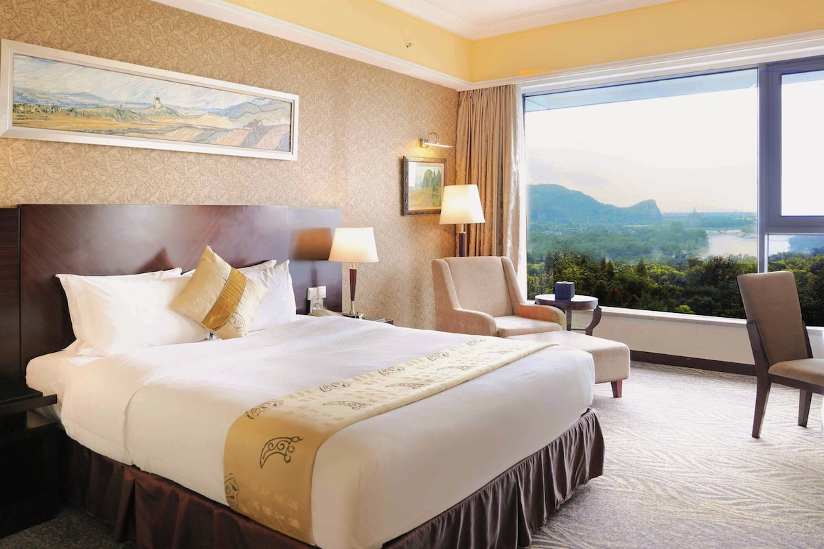 Quarto de hotel com uma cama grande, duas cadeiras, uma mesa de cabeceira e vista para uma paisagem através de uma janela ampla.