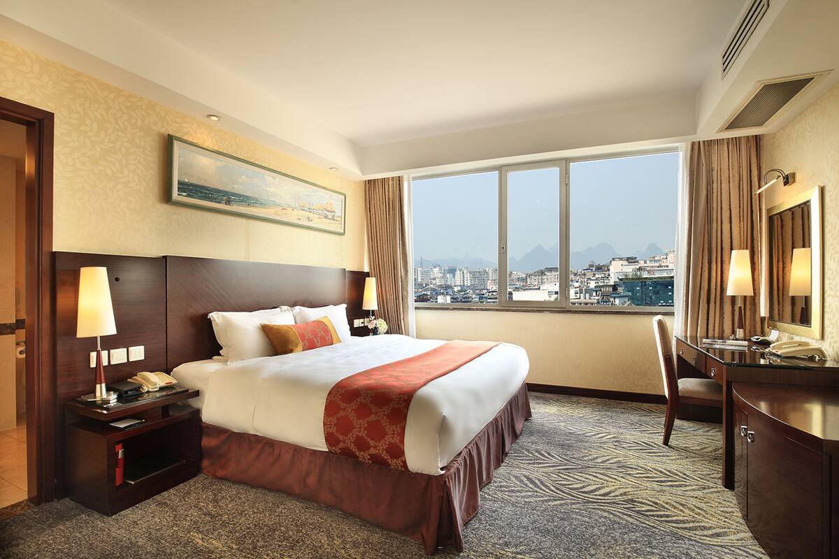 Quarto de hotel com uma cama grande, uma escrivaninha com cadeira, uma janela com vista para a cidade e um quadro na parede.