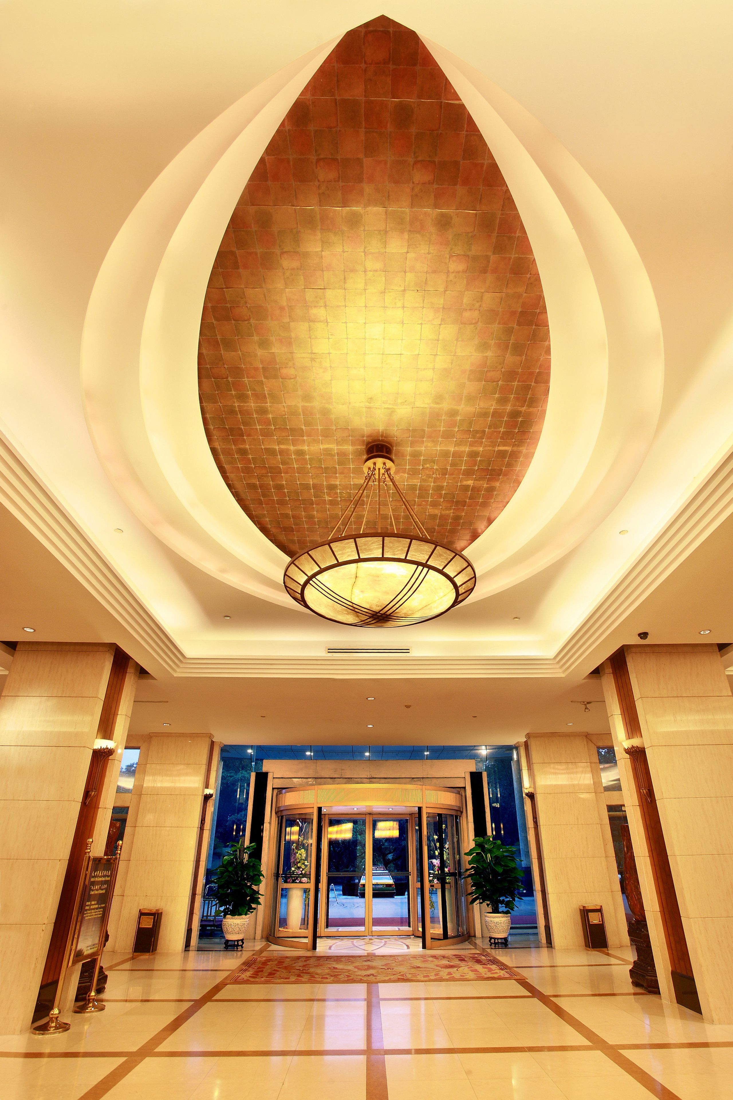 A hotel lobby with a large, arched ceiling and a central chandelier. There are potted plants and a reception desk in the foreground.