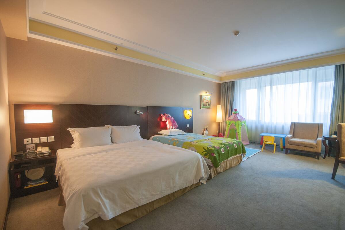 Quarto de hotel com duas camas, uma escrivaninha, uma cadeira e uma janela grande com cortinas.