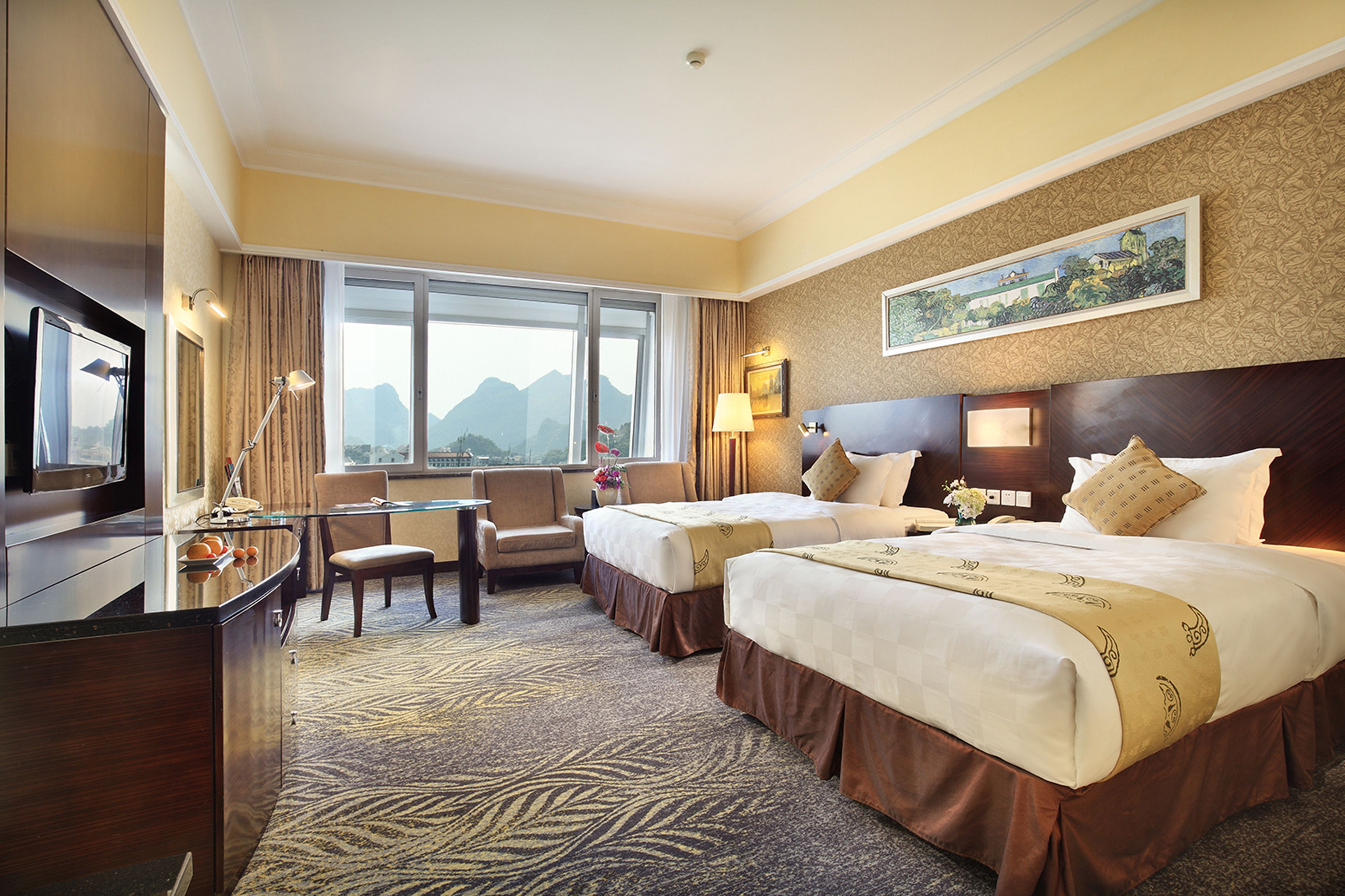 A hotel room with two beds, a desk, a chair, and a large window with a mountain view.