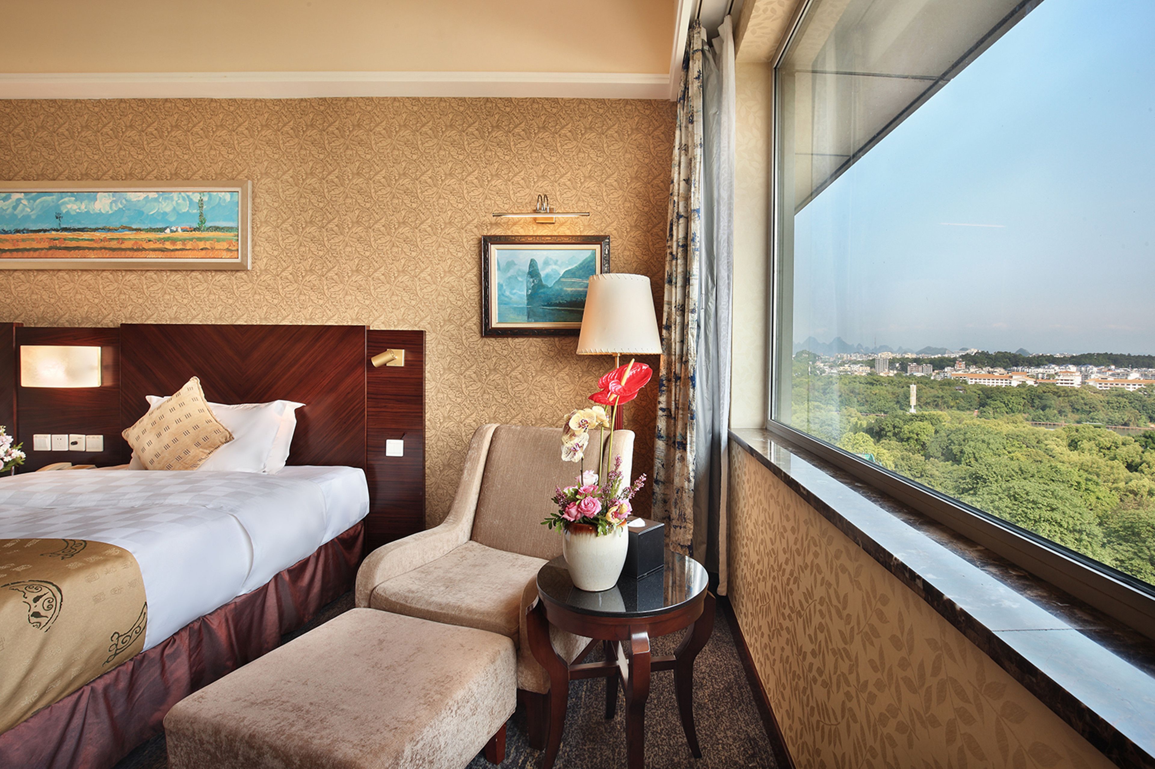 Habitación de hotel con una cama grande, una silla, una mesita con un florero y una lámpara, además de un cuadro en la pared.