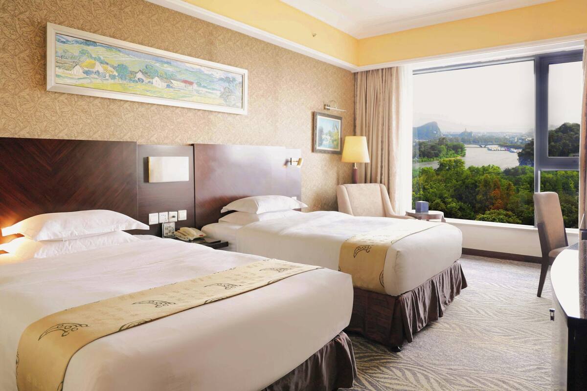 Quarto de hotel com duas camas, uma janela grande com vista para um rio e paisagem urbana, um quadro na parede e uma escrivaninha com abajur.