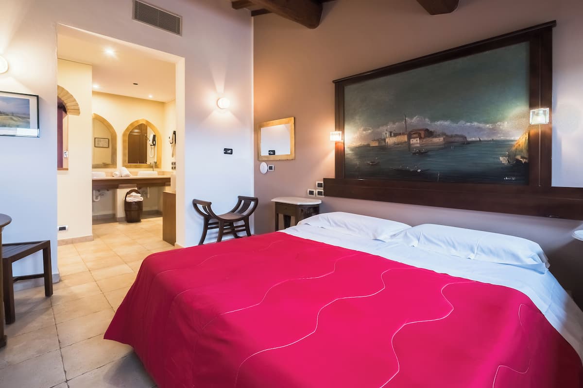 Una habitación de hotel con una cama grande, un escritorio, una silla, un baño con lavamanos y espejo, y un cuadro en la pared.