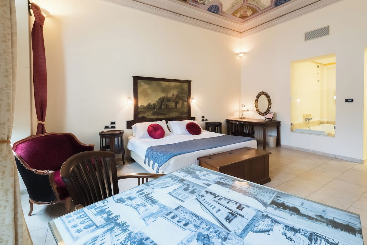 Habitación de hotel con cama, escritorio, silla, un cuadro y baño visible a través de una puerta de cristal.