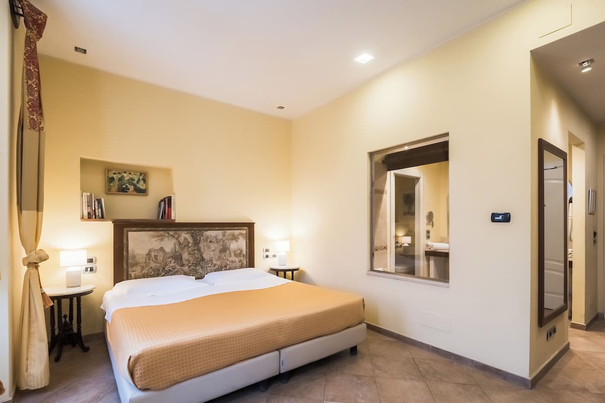 Una habitación de hotel con cama, mesitas de noche, un cuadro, una ventana y una puerta.