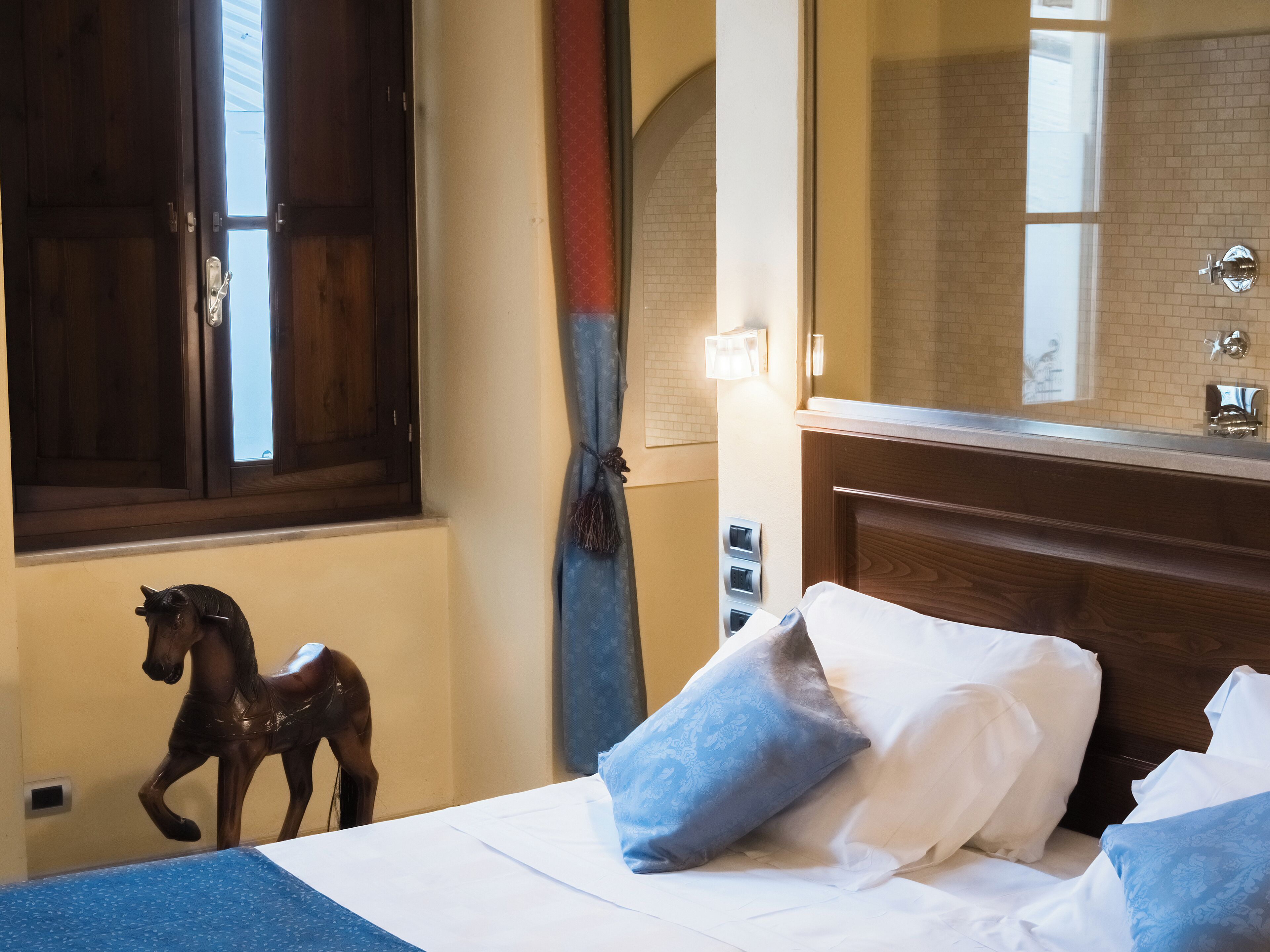 A hotel room with a wooden headboard, a bed with white and blue pillows, a small horse statue, a window with a view of a brick wall, and a mirror above the sink.