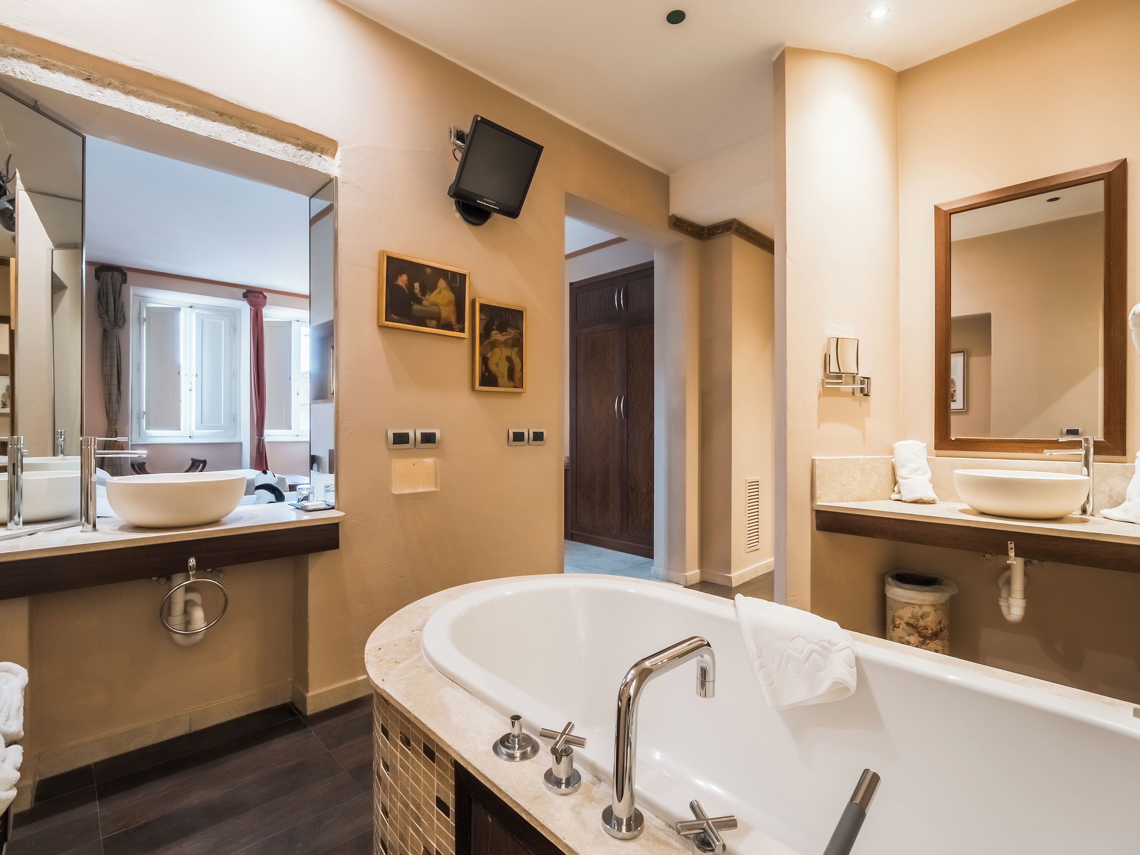 A modern bathroom with a large bathtub, two sinks, and a flat-screen TV mounted on the wall.