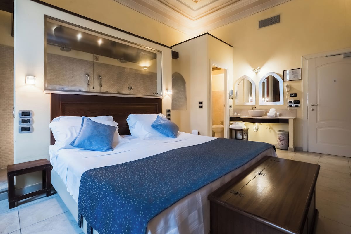 Habitación de hotel con una cama grande, una mesita de noche, un escritorio, un tocador con espejo y un baño con lavamanos e inodoro.