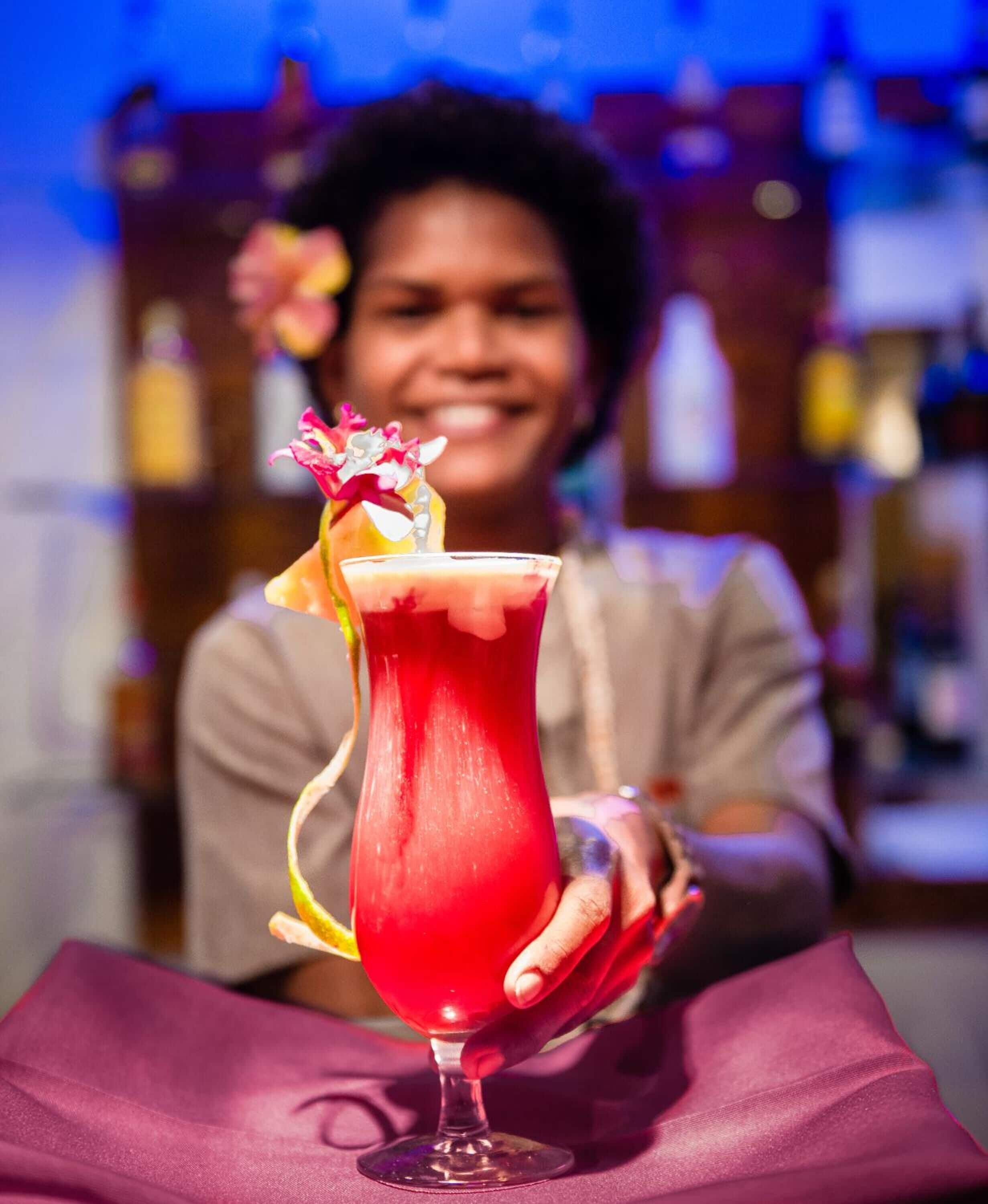 A person seated at a table with a vibrant red drink garnished with a flower, in a setting with a blurred background suggesting a bar or lounge area.