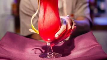 A person seated at a table with a vibrant red drink garnished with a flower, in a setting with a blurred background suggesting a bar or lounge area.